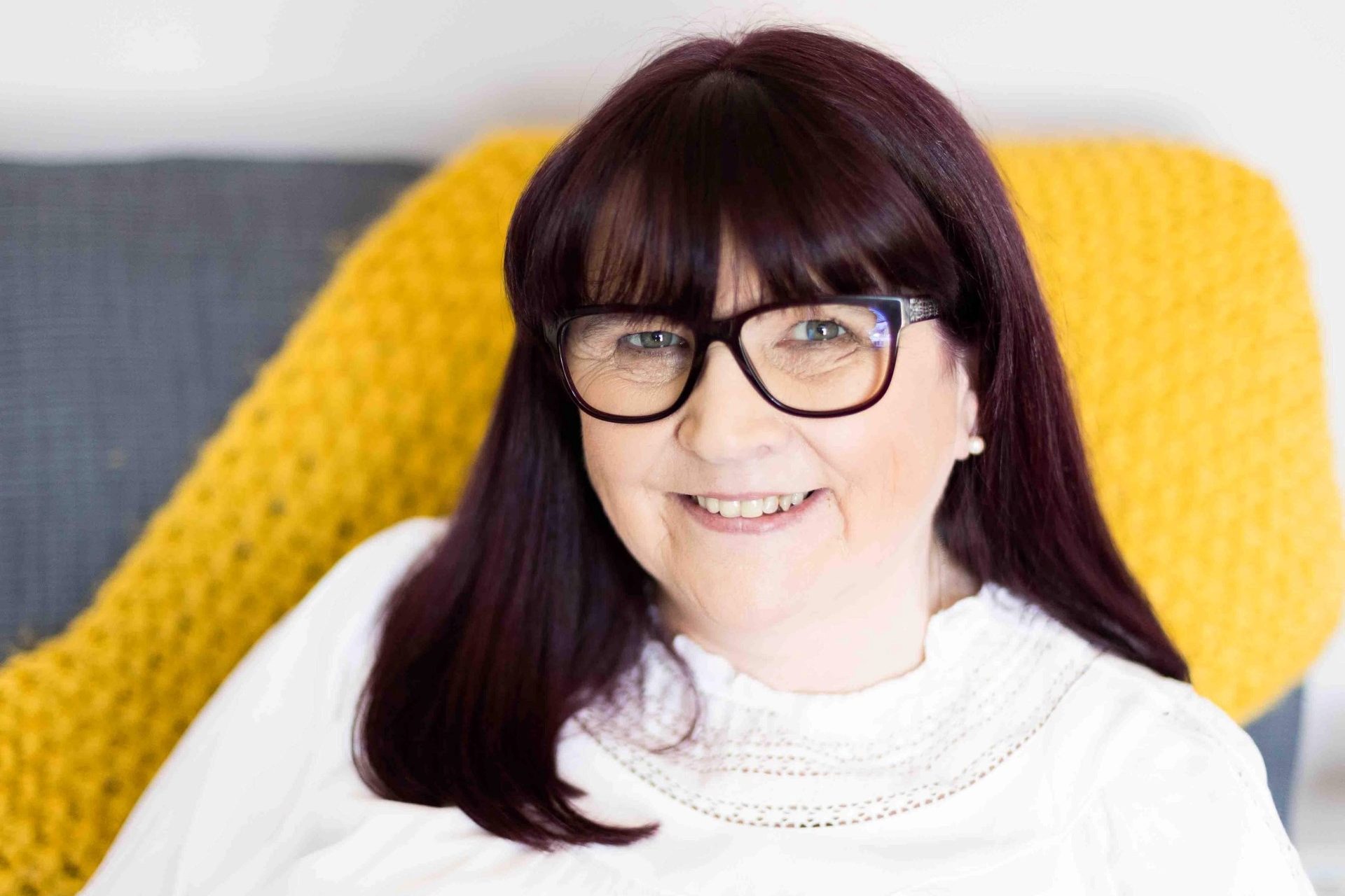 Image a a women with long dark hair wearing a white top. Sitting on a grey sofa with a mustard colour throw on.